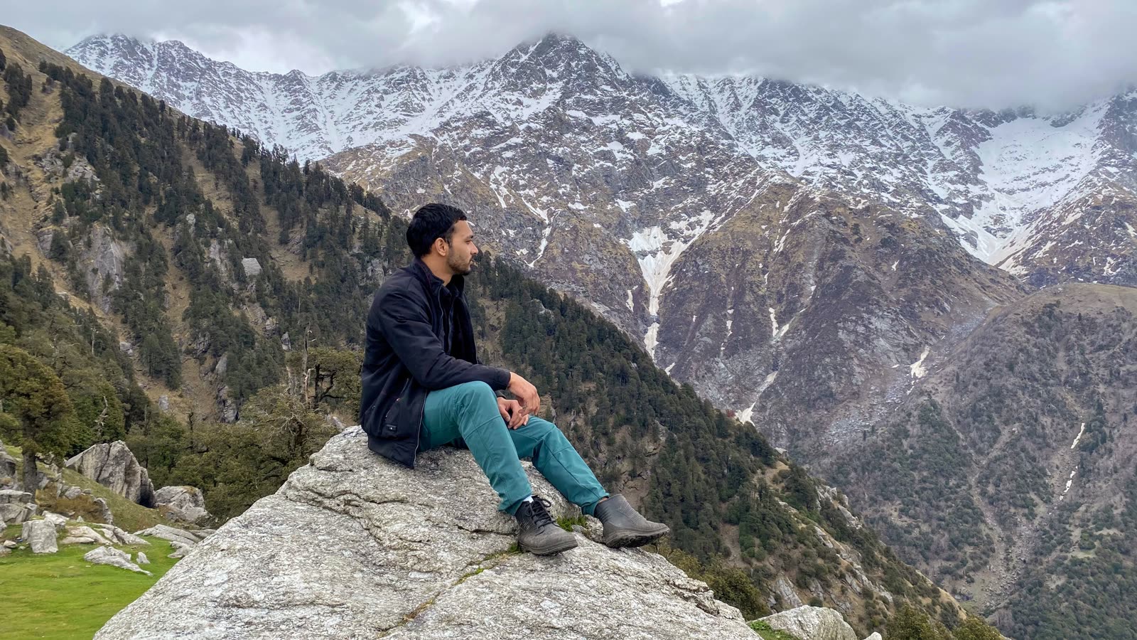 Saurav sitting on a big rock, looking out at snow covered mountains under clouds